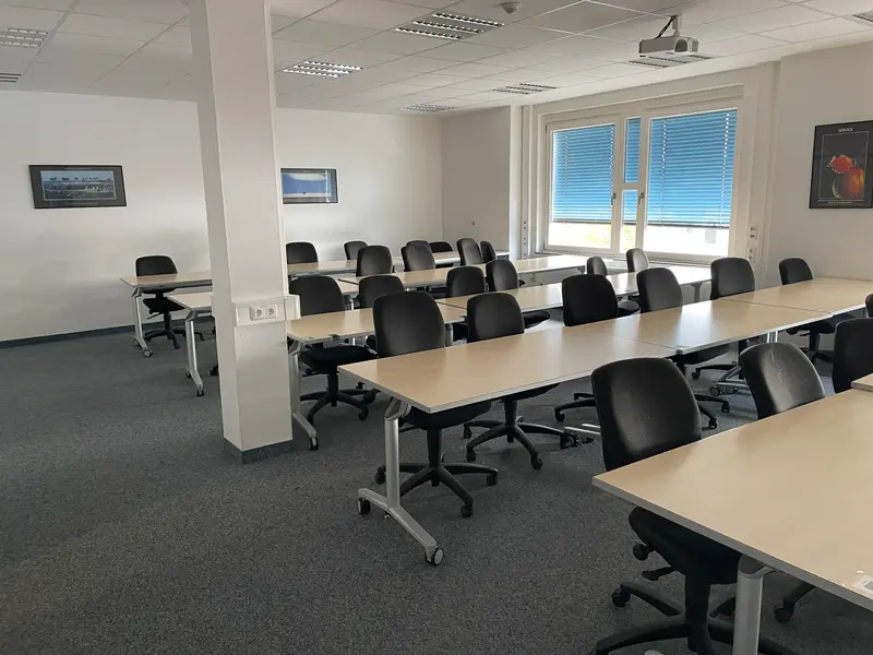 A brightly lit lecture room at HSW University of Applied Sciences with beige wooden tables and black wheelchairs on gray carpet, a projector on the ceiling and pictures with motivational sayings on the walls.